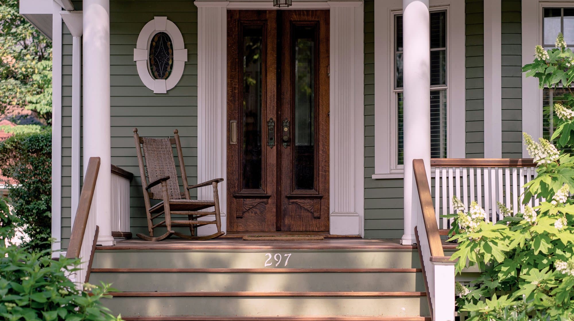 A green-clapboard front porch with a wooden rocking chair and dark double doors.