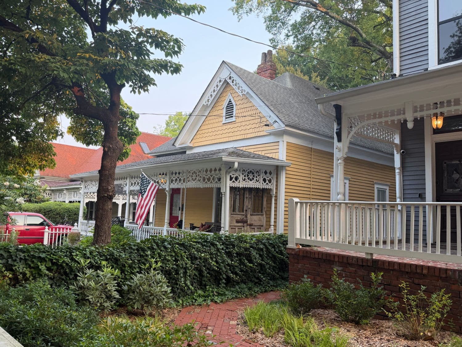 A row of older Atlanta houses with deep front porches, including a yellow Victorian with white gingerbread trim and an American flag, behind an ivy-covered wall and a brick walk.