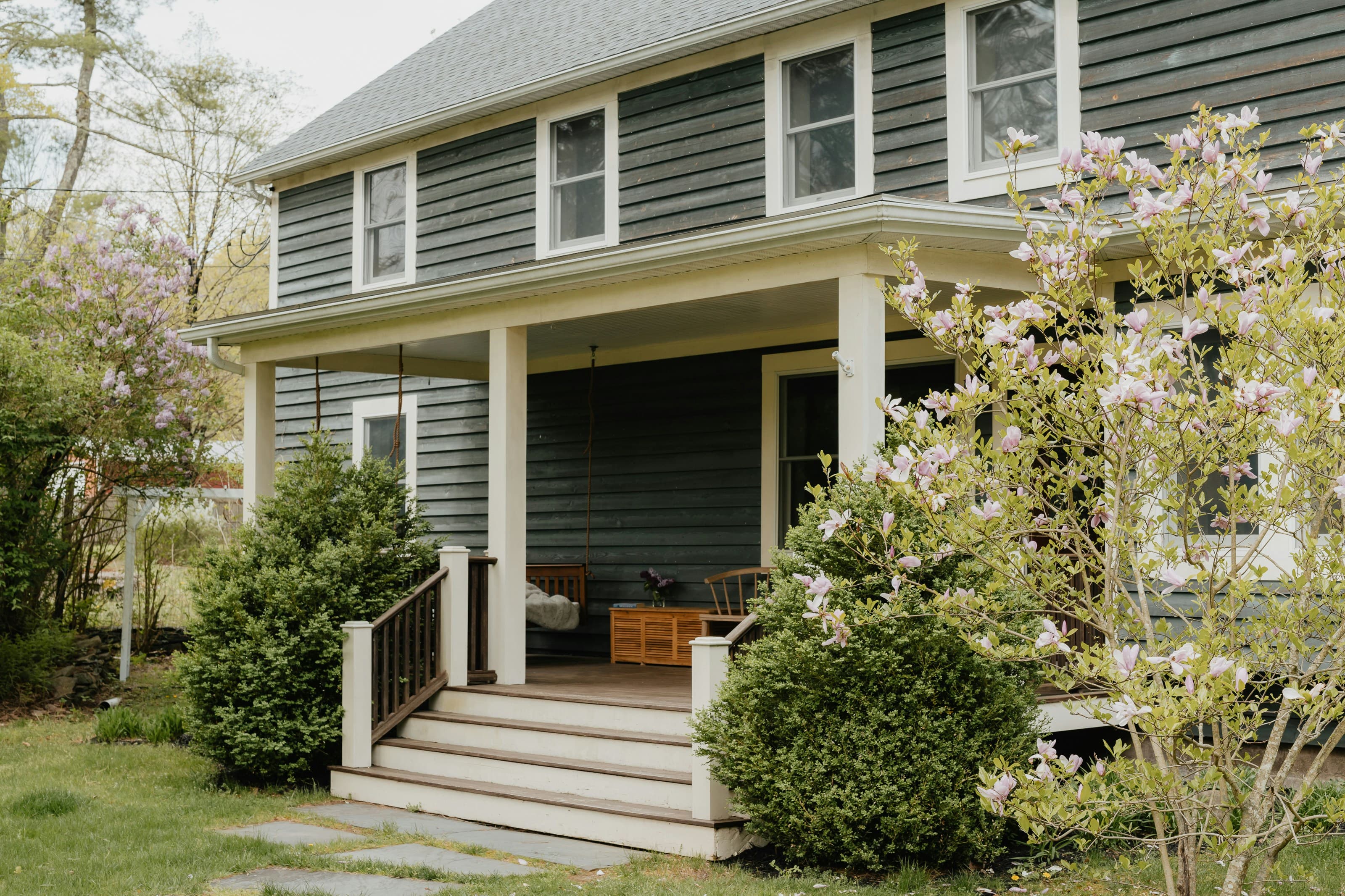 A modest two-story home with dark green wood siding and a covered front porch, framed by flowering shrubs.